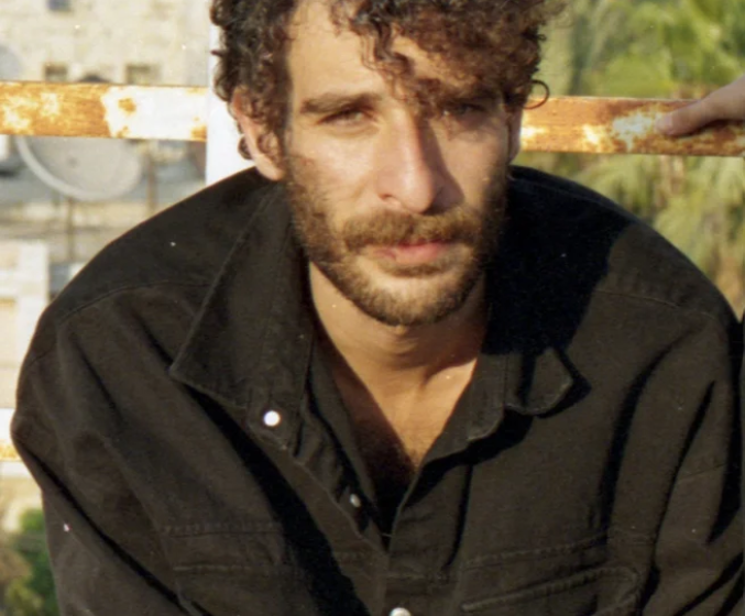 Close-up portrait of a man with curly hair and a beard, wearing a black shirt, sitting against a rustic background.