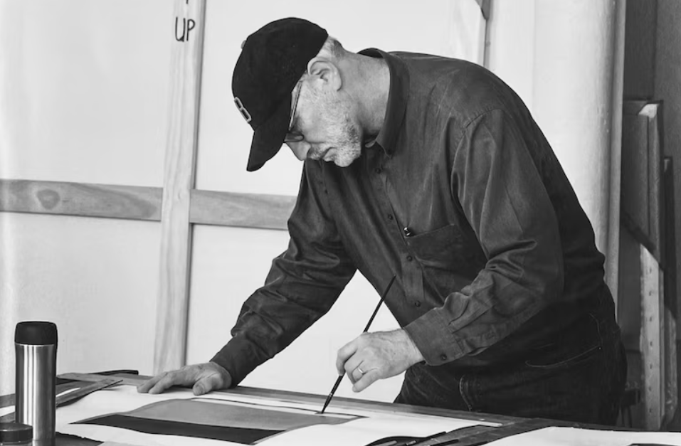 A man with glasses and a black cap is painting on a large sheet of paper in a studio, focused on his work.