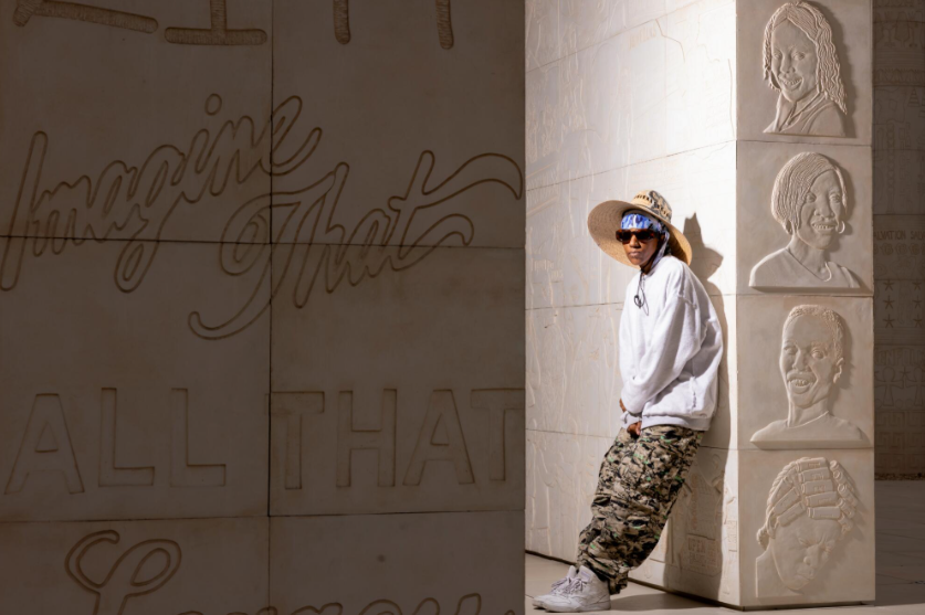 A person wearing a white hoodie, sunglasses, and a straw hat stands next to a textured wall featuring engraved portraits and text that reads 'Imagine That' and 'ALL THAT'.