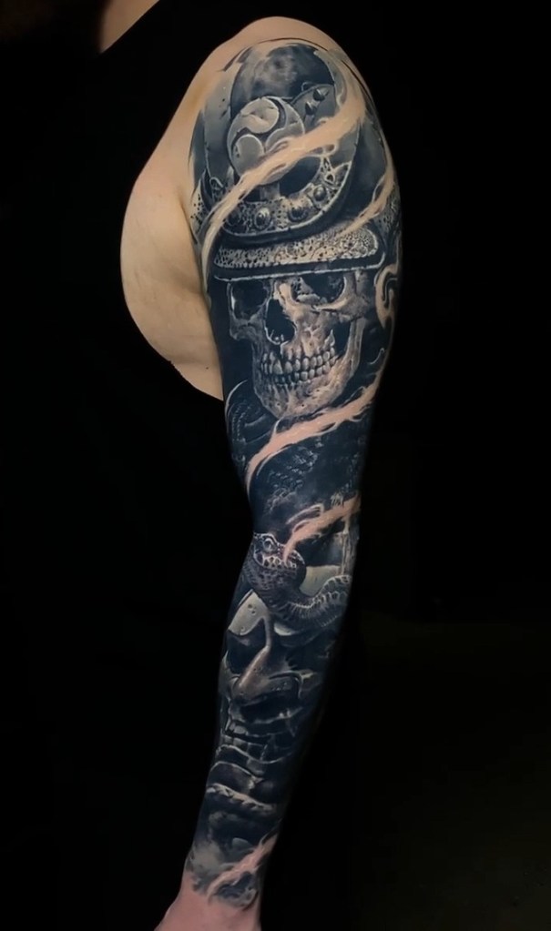 A detailed tattoo on a man's arm featuring a skull, samurai helmet, and swirling clouds, with dark shading and vibrant colors.