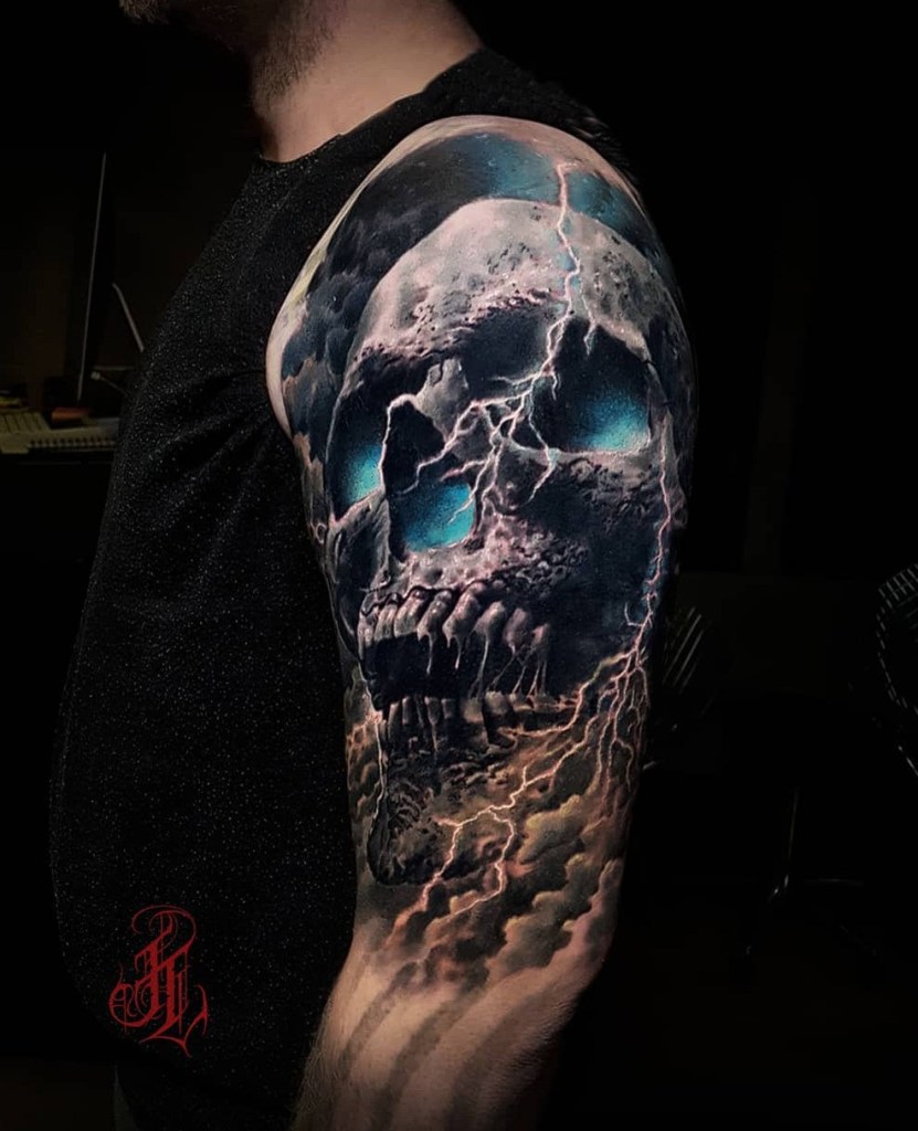 A close-up of a detailed tattoo featuring a skull with glowing blue eyes, surrounded by storm clouds and lightning, on a person's upper arm.