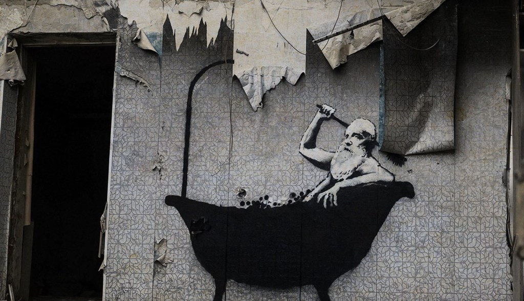 Street art mural depicting a figure with a beard bathing in a bathtub, set against a dilapidated wall with peeling paint.