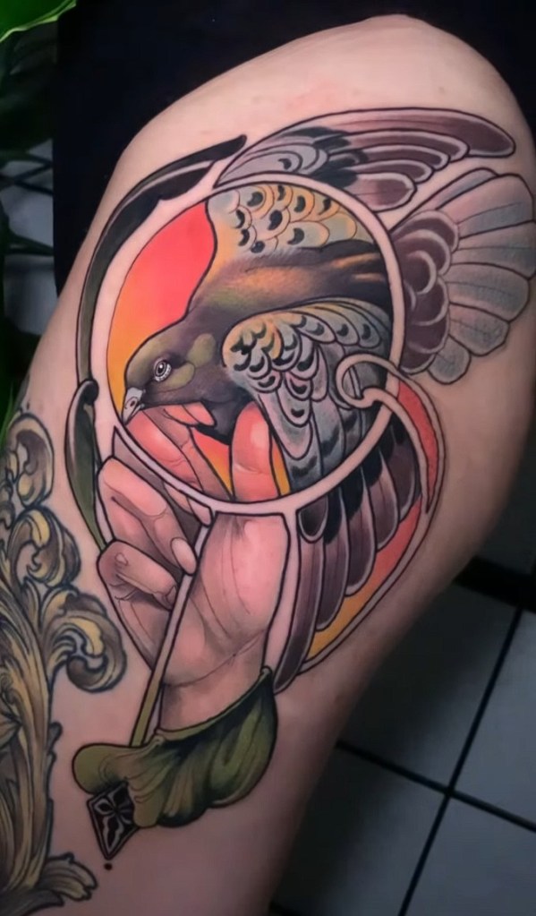 A colorful tattoo depicting a bird in flight, entangled in a circular design, with a hand gripping a decorative element.