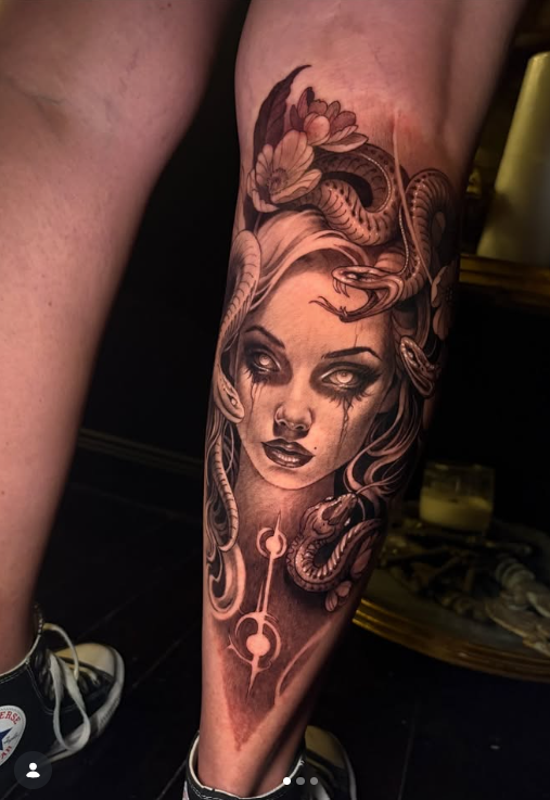 A detailed tattoo design on a leg featuring a woman's face with long hair, adorned with snakes and flowers, showcasing intricate shading and facial expressions.