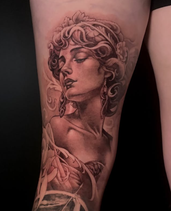A detailed tattoo on a thigh depicting a woman with curly hair and elegant accessories, styled in a vintage manner, with flowing lines and soft shading in a monochromatic color scheme.