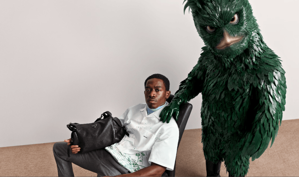 A young man seated in a chair holding a black handbag, with a green feathered figure resembling a bird standing behind him.