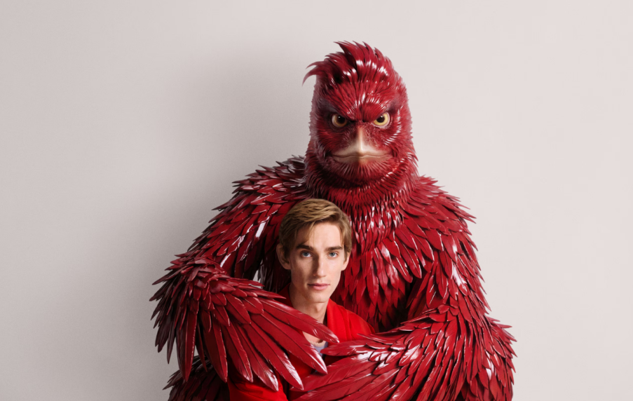 A young man poses beside a large, anthropomorphic red bird character with intricate feather details, both against a plain background.