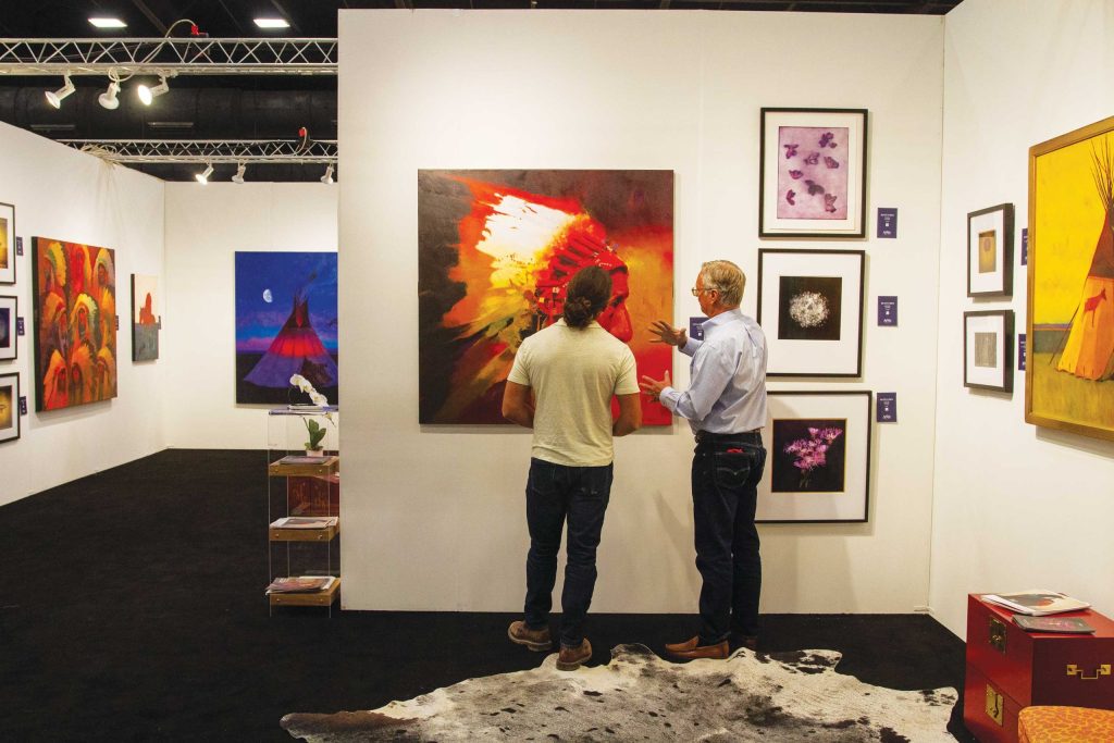 Two men discussing artwork in an art gallery with various paintings displayed on the walls.