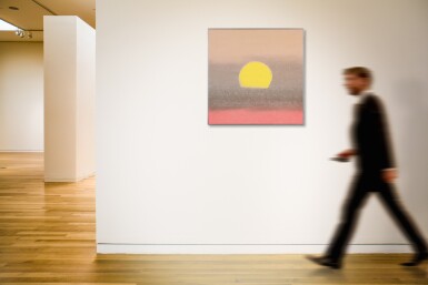 A blurred figure of a man walking past an abstract painting of a sun on a gallery wall.