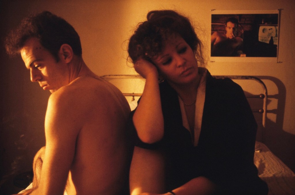 A man and woman sit back to back on a bed in a dimly lit room, expressing emotional distance. The woman appears contemplative, resting her head on her hand, while the man looks away. A photograph of a man hangs on the wall in the background.