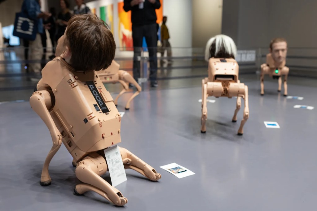 A group of humanoid robots with heads resembling human faces, positioned on a gallery floor among scattered cards, showcasing an installation art piece.