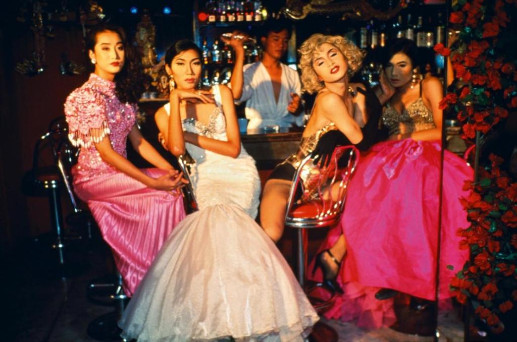 Four individuals dressed in glamorous attire sitting at a bar, showcasing fashion styles including a sequined dress and pink skirts, with a bartender in the background.