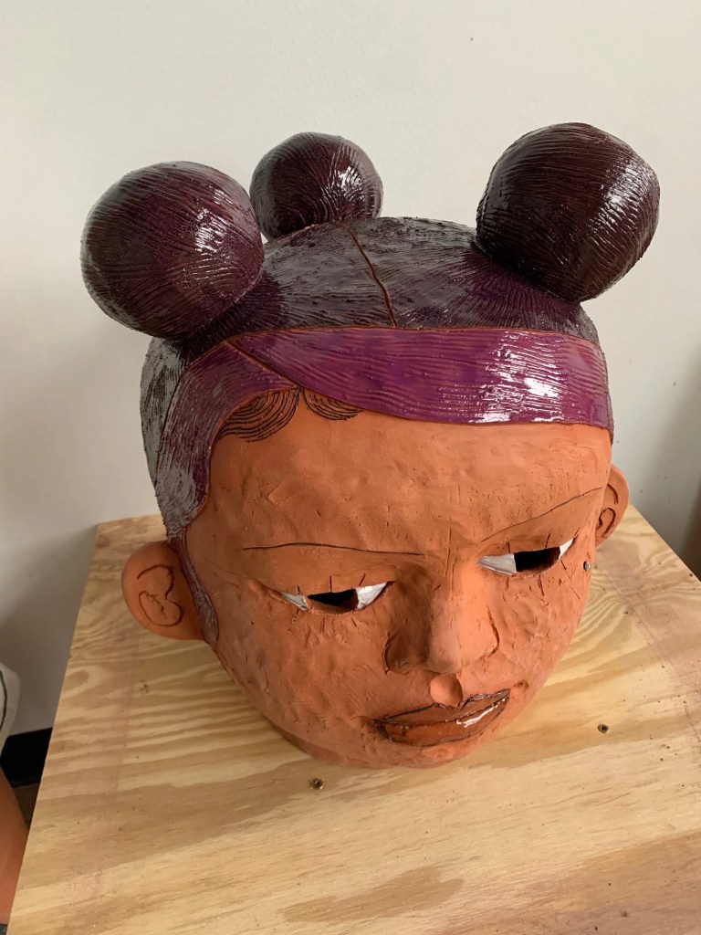 A clay sculpture of a stylized human head with two rounded hair buns and a serious expression, displayed on a wooden surface.