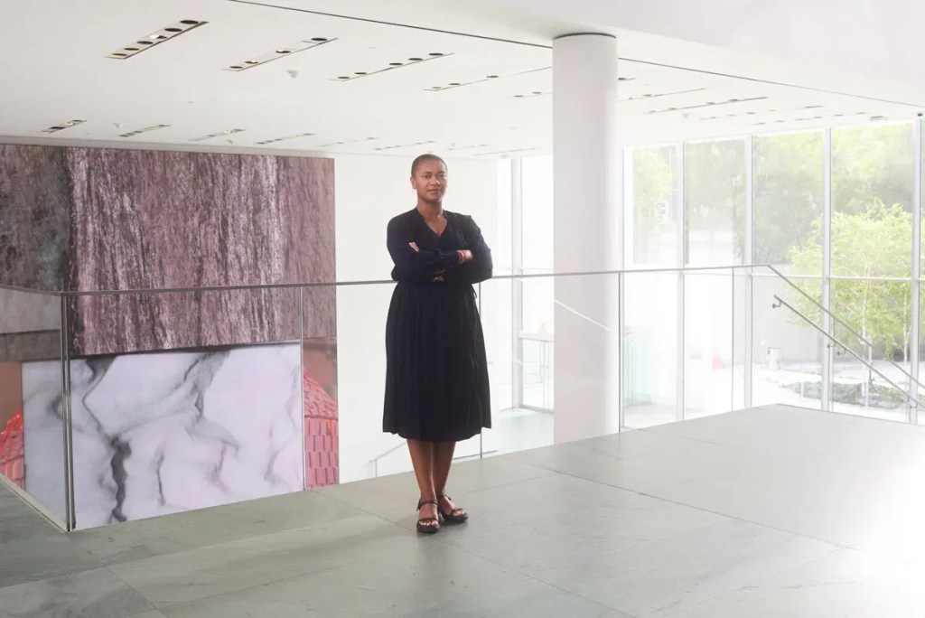 A person stands confidently with arms crossed in an art gallery, featuring modern architecture and colorful backgrounds.