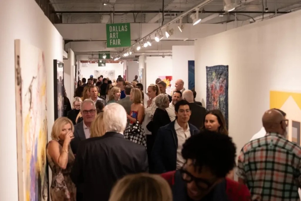 A crowded art gallery during the Dallas Art Fair, with attendees admiring various artworks displayed on the walls.