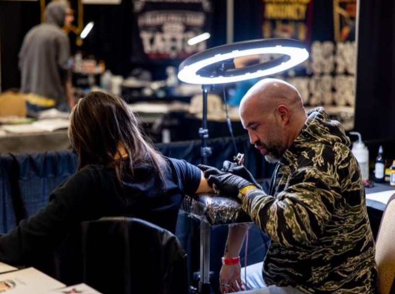A tattoo artist working on a client's arm under a circular light, with additional tattooing supplies visible in a convention setting.