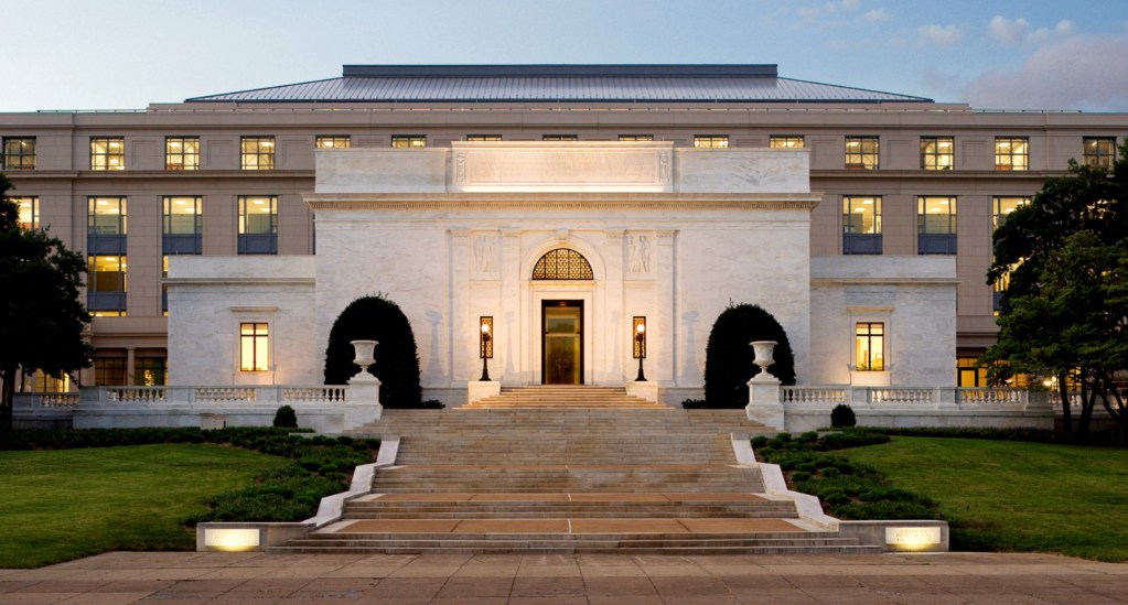 Exterior view of a grand building featuring marble columns, large steps, and illuminated windows, set in a landscaped area.