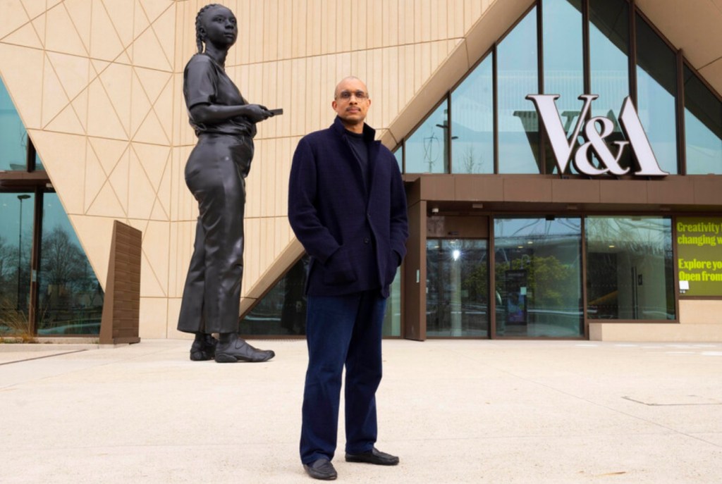 A person stands in front of the V&A Museum, with a large black statue of a woman behind them. The statue is holding a phone and wearing a casual outfit, showcasing a modern architectural backdrop.