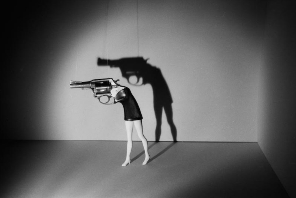 A stylized black and white image depicting a mannequin holding a toy gun, with its shadow cast on the wall, creating a dramatic effect.