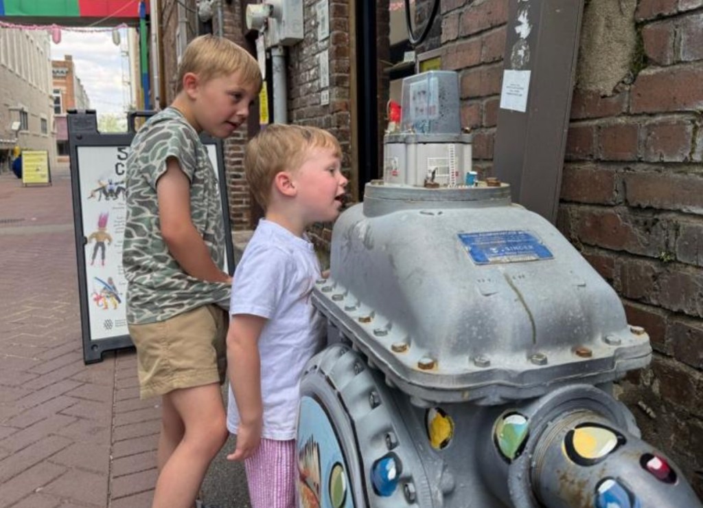 Two young boys observing a colorful machine in an urban setting, with a signboard in the background.