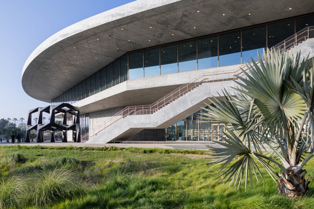 Modern architectural building with large glass windows and an oval roof, featuring outdoor stairs and a contemporary art sculpture in front, surrounded by greenery.