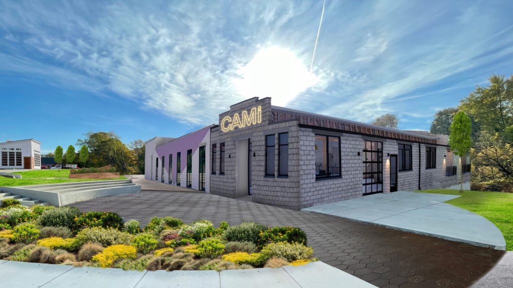 Exterior view of a modern building with a sign reading 'CAMI,' surrounded by landscaped gardens and a clear blue sky.