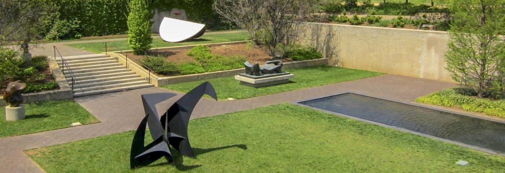 An artistic outdoor sculpture garden featuring abstract sculptures, manicured lawns, a water feature, and walking paths surrounded by trees.