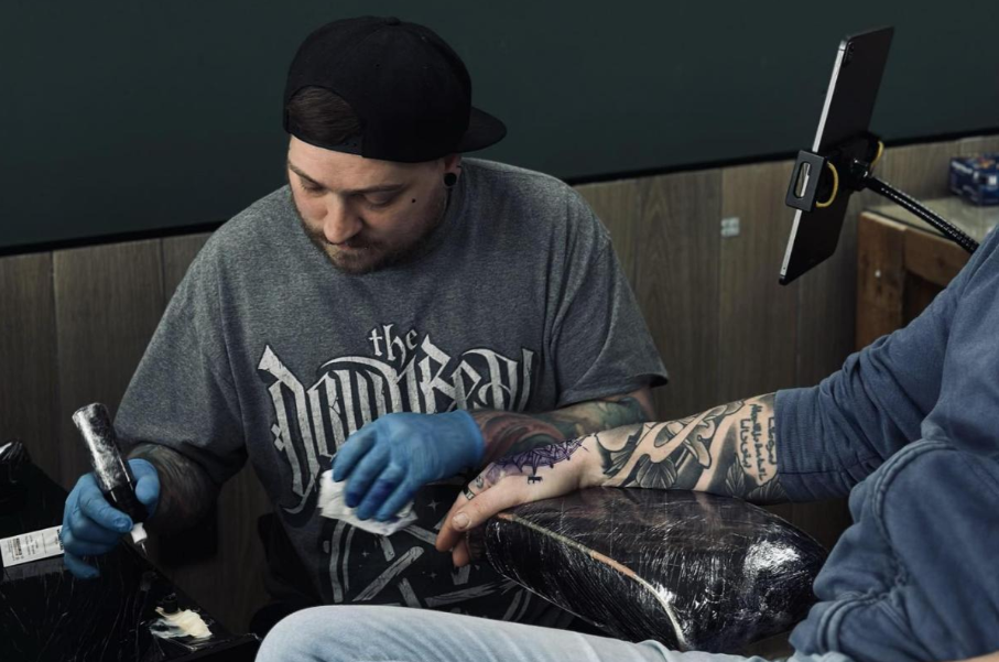 A tattoo artist with gloves is applying ink to a client's arm, which is covered in tattoos, while using a machine. The artist wears a grey t-shirt and a cap, focused on the task.