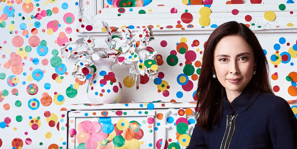 A woman with long dark hair sits in a colorful room decorated with paper dot patterns on the walls and furniture, and a white vase with paper flowers.