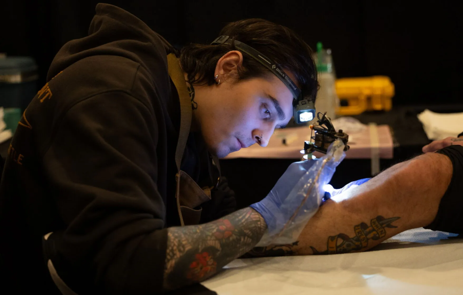 A tattoo artist focused on inking a design on a client's arm, using a tattoo machine and wearing gloves. The artist has visible tattoos and a headlamp for better visibility.