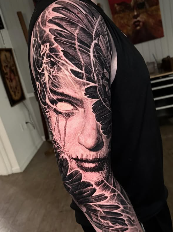 A close-up of a detailed tattoo on a person's arm, showcasing a face with striking features amidst intricate black feather designs, conveying a haunting aesthetic.