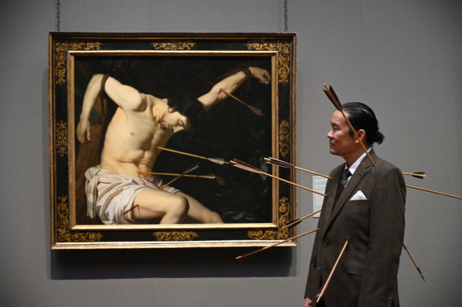 A man dressed in a suit stands beside a painting of a figure pierced with arrows, resembling a dramatic scene of suffering, in an art gallery setting.