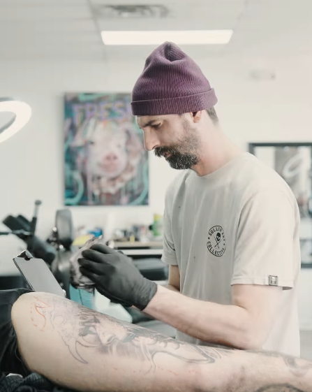 A tattoo artist wearing a beanie and gloves works on a client's leg tattoo in a studio, with art on the walls in the background.