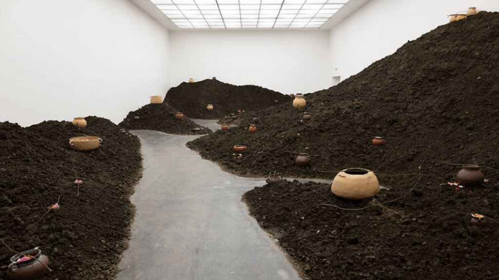 An art installation featuring mounds of dark soil with clay pots scattered throughout and a pathway running through the center.