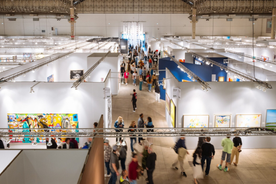 A spacious art exhibition with numerous visitors exploring various artworks displayed on white walls.
