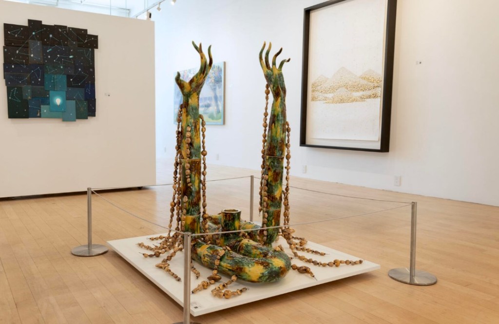 A sculptural installation featuring organic forms resembling elongated hands, adorned with beads, displayed on a pedestal in a gallery space. In the background, abstract artworks and framed pieces hang on the walls.