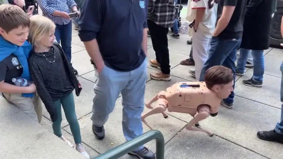 A robotic dog-like device featuring a human head moves through a crowd, with children appearing amused nearby.