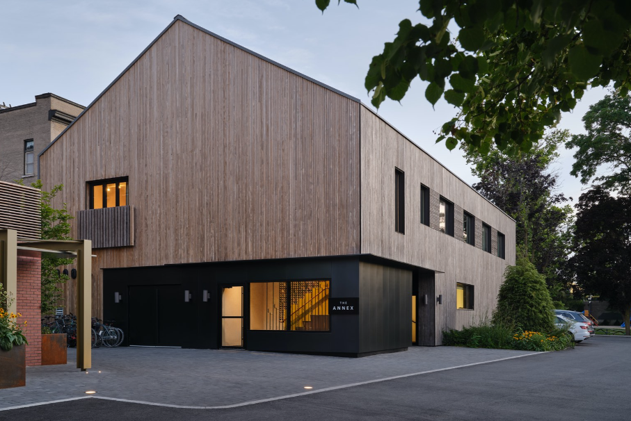 A modern wooden building with a sloped roof and large windows, featuring a dark entrance labeled 'THE ANNEX'. The surrounding area includes greenery and parked bicycles.
