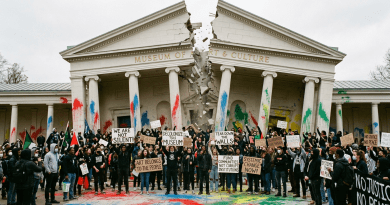 Protesters holding signs in front of a museum with paint-splattered columns and a cracked facade