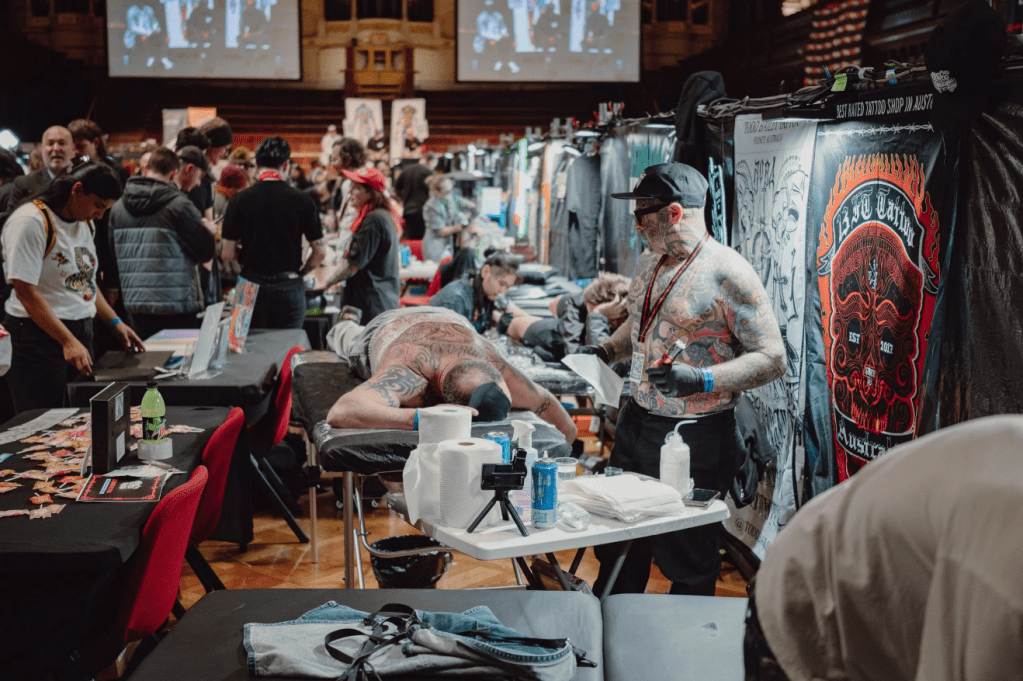 Vibrant scene from a tattoo expo with artists working on clients amid a crowded convention hall filled with attendees and vendor booths.

