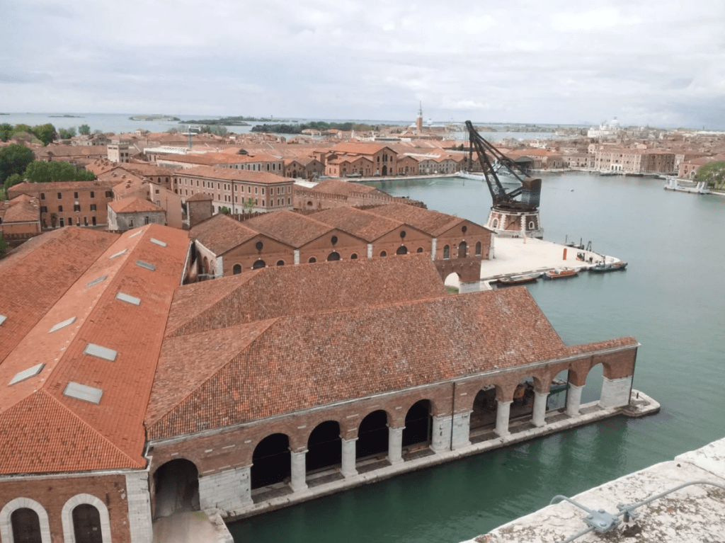 Aerial perspective of Venice's Arsenale complex with historic brick buildings, arched structures, and waterfront, a primary exhibition site for the 2026 Venice Biennale.


