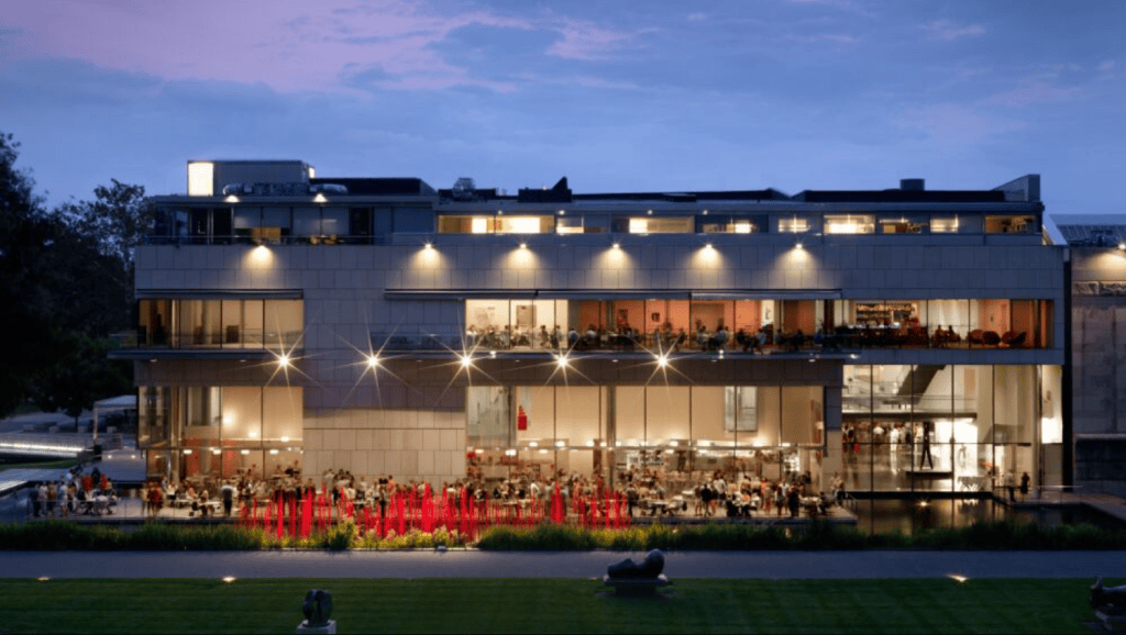 Exterior view of the Virginia Museum of Fine Arts at twilight, showing its modern glass and stone architecture with warm interior lighting and visitors gathered outside.

