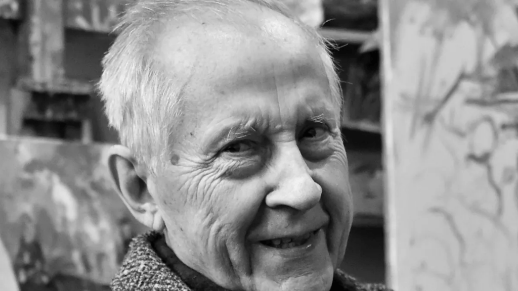 Black-and-white close-up portrait of elderly German painter Harald Metzkes smiling slightly, with white hair and studio background, 2014 or later.  

