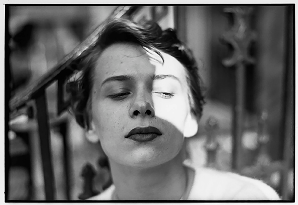 Close-up black and white photographic portrait of a woman with strong contrast lighting casting a shadow across half her face, highlighting expressive eyes and emotional intensity.

