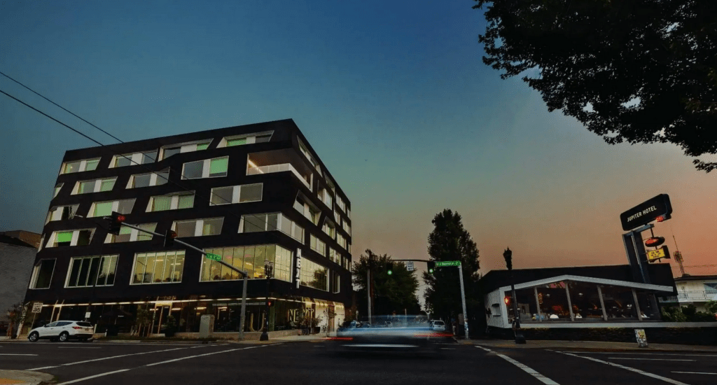 Exterior view of the modern black Jupiter NEXT building at dusk with the Jupiter Hotel sign visible, located in Portland, Oregon.  


