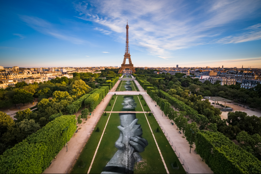 Aerial view of massive land art painting of giant hands clasped together on green lawn with Eiffel Tower visible.