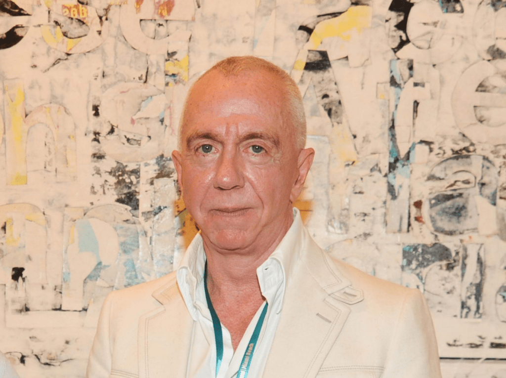 Professional headshot of Brent Sikkema, an older man with short light hair wearing a white jacket, standing in front of abstract art.
