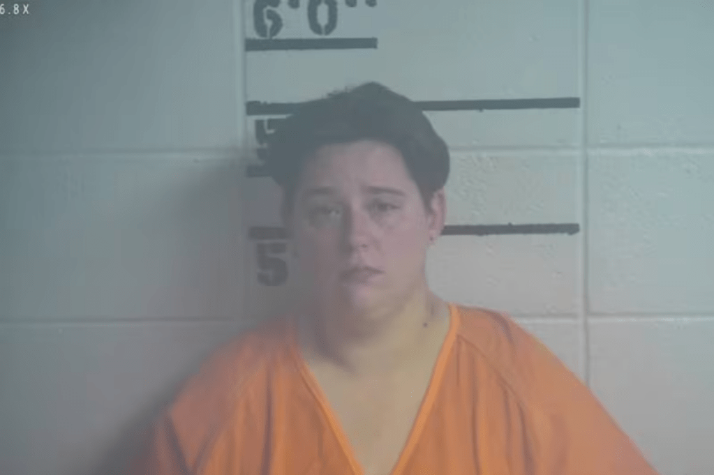 Mugshot of Brook McDaniel, a woman with short dark hair wearing an orange jail uniform, standing against a height chart background.