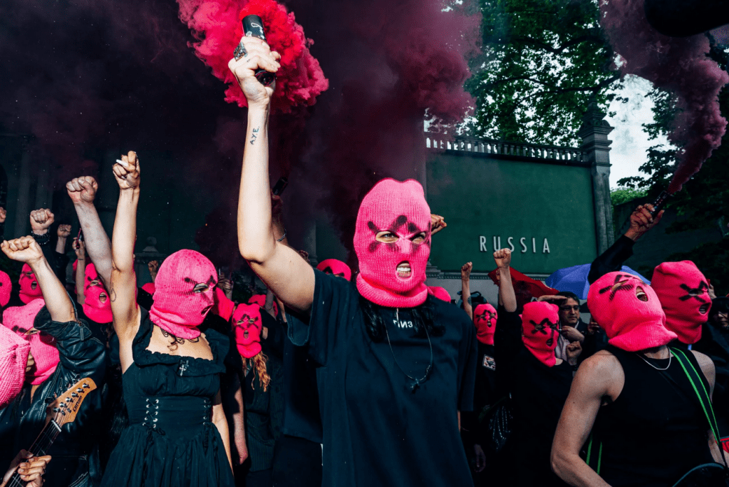 Group of activists in pink balaclavas and black clothing raising fists and smoke flares in front of the green Russian Pavilion building, with "RUSSIA" visible.

