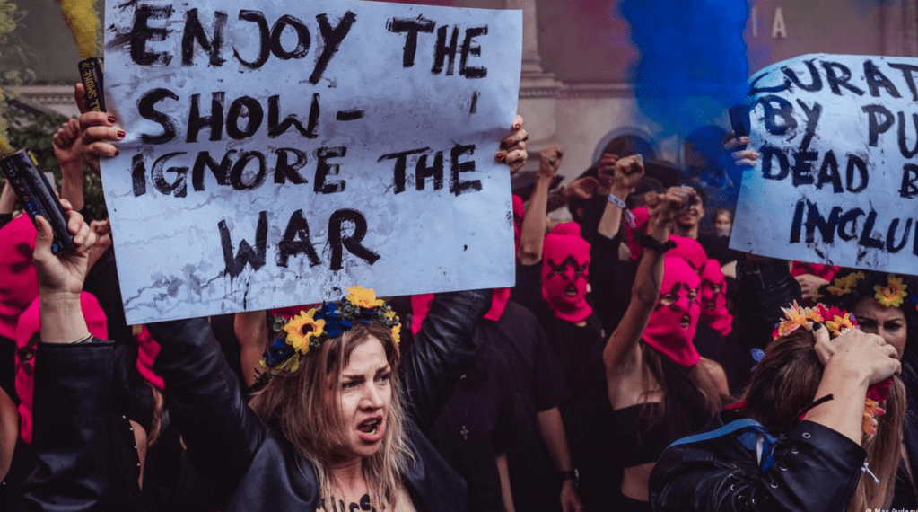 Demonstrators holding signs with messages like “Enjoy the Show – Ignore the War” during Biennale-related protests, with smoke and crowds visible.

